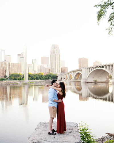 Couple kissing on a rock near the Stone Arch Bridge in Minneapolis, with the Mississippi River and city skyline in the background. Bride wears a stunning long red dress. Captured by Toly Dzyuba Photography, Minneapolis wedding photographer.