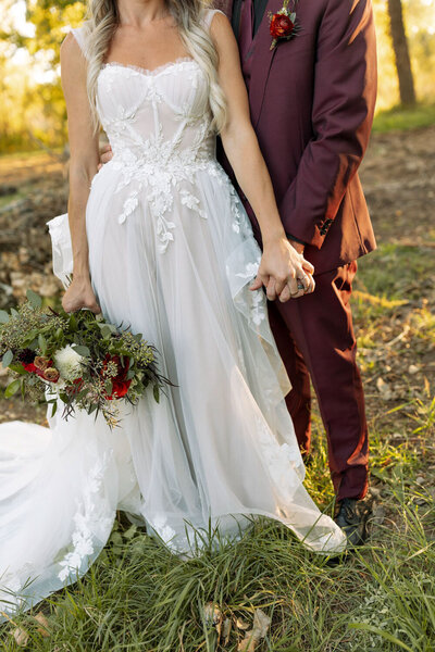 Close-up of a couple holding hands, bride holding a bouquet, groom in a burgundy suit