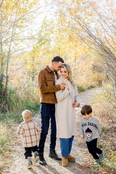 Mom and dad standing together as two little children run around them in a Calgary, AB park.