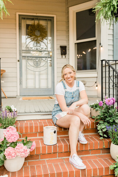 A DIYer sitting on the front step of her old  victorian home, with a paint can next to her. Potted flowers on the front porch surround either side of the steps.
