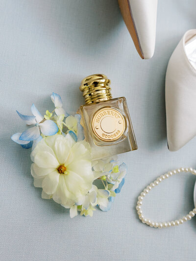 Bridal detail flat lay with burberry perfume bottle, white ranunculus, and blue hydrangeas and pearl accents