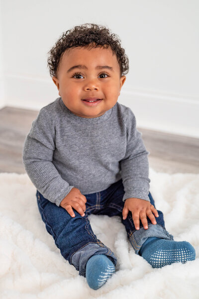 A baby boy sits on a blanket and smiles at the camera for his nine month pictures