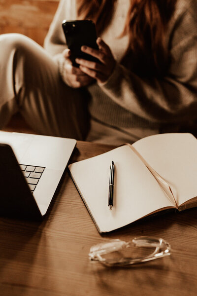 kvinna sitter med mobil i handen och har dator och anteckningsbok framför sig