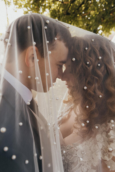 Bride and groom embrace underneath pearl-adorned wedding veil
