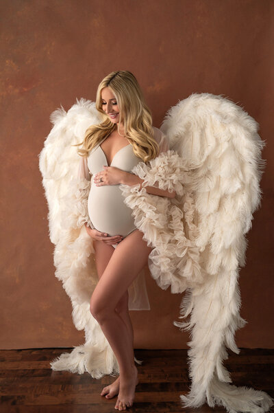 Expecting mother wearing a simple, nude-toned bodysuit, posing with angel wings with soft studio lighting in Waukesha, Wisconsin.