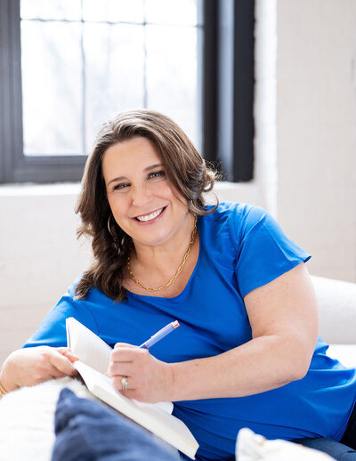 Chris smiling while writing in a notebook, sitting on a white couch in a bright room, wearing a blue blouse.