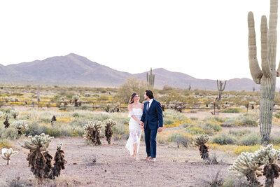 Bride and groom walking through the Arizona desert holding hands with mountains and cacti in the background.