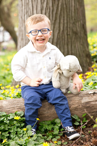 Little boy sits on log in yellow flowers with stuffed animal smiling at camera
