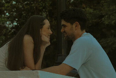 A couple are across from each other at a picnic table. The woman is leaning closer to the man with her elbows on the table and her head resting on her hands.