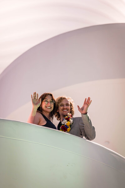 A couple on their elopement day at the Moss Arts Center look down and smile as they wave from the second floor.