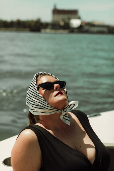 Woman in striped headscarf and black swimsuit basks in the sun on a boat with the St. Augustine waterfront in the background.