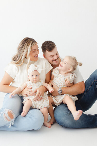 A family of four sitting on the floor in a photography studio looking at each other and laughing.