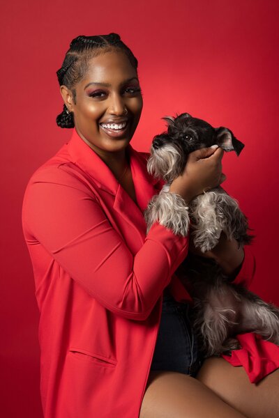 Indoor portrait of lady wearing a red blazer on a red backdrop with her miniature schnauzer in hand.