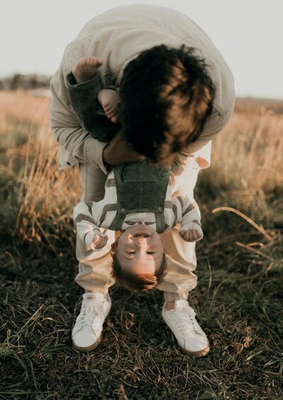 Man standing in field with his toddler son, bending over and holding the toddler upside down. 