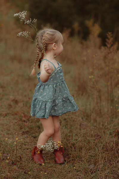 Little girl in denim dress dances in the tall grass with wildflowers by Lawrenceville Children's Photographer