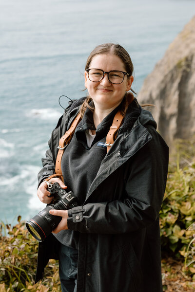 Desiree standing and holding her camera. She is on a cliff edge with the ocean behind her.