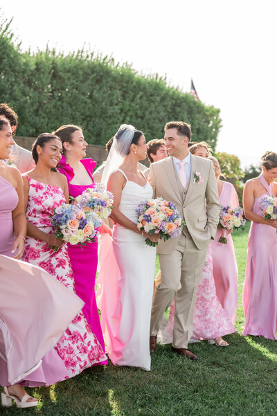 A joyful bridal party in vibrant pink and floral gowns surrounds the bride and groom during their Cape Cod wedding at Wychmere Beach Club. The perfect mix of coastal elegance and playful style.