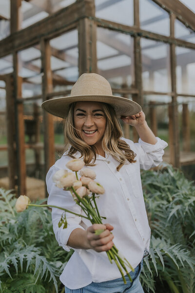 Alexandra holding up flower stems and smiling at the camera, one hand on her hat