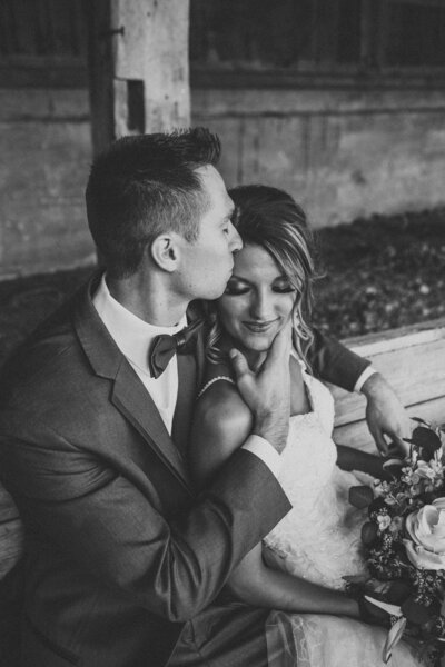 Black and white photo of groom sitting down with groom pulling in brides cheek for a kiss by Iowa City wedding photographer Sabrina Wilham