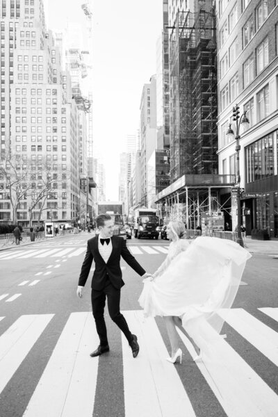 Black and white photo of a bride and groom crossing a city street hand-in-hand, with the bride’s dress flowing behind her amid tall skyscrapers.