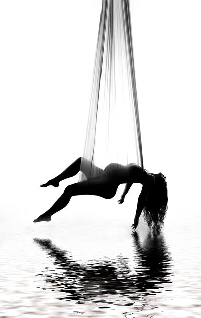 Luxury studio maternity photography session at top Philadelphia, PA studio - Mom is suspended from silks from the ceiling with her legs and arms stretched toward the ground. There is the illusion she is suspended over water with her reflection in the water.
