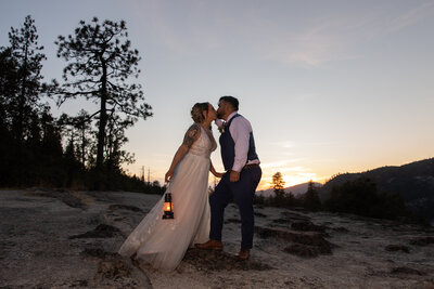 A bride wearing a white dress and holding a lantern at her side leans in to kiss her groom.