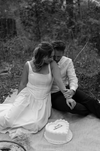 Bride and groom sitting on a blanket with their wedding cake, in black and white.