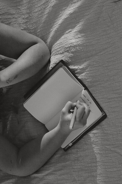 A black and white photo of a woman writing in her journal laying down with her head propped up by her hand