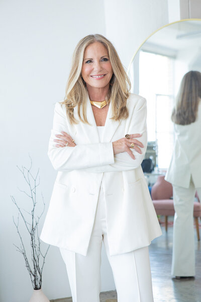 woman in a white suit smiling with arms crossed standing in a modern room with a mirror reflecting her image