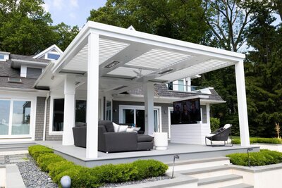 White StruXure pergola with white louvers on a raised platform with landscaping and furniture. 