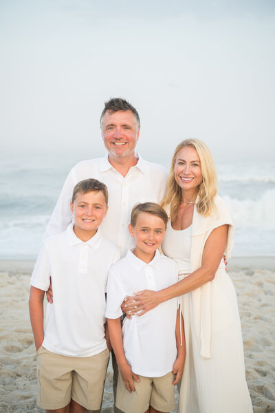 a family dressed in white and khaki on the beach standing together and smiling with a light foggy haze surrounding them with the ocean in the background