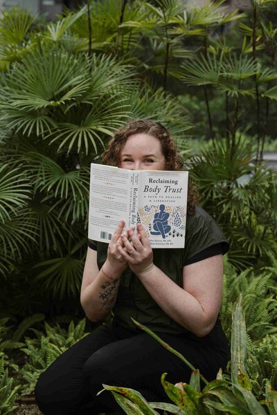 Christy Walowit sits outdoors, partially hiding her face with the book "Reclaiming Body Trust." She's surrounded by lush green plants, creating a peaceful atmosphere.