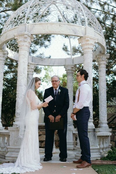 A bride reads her vows to her groom in front of a pergola.