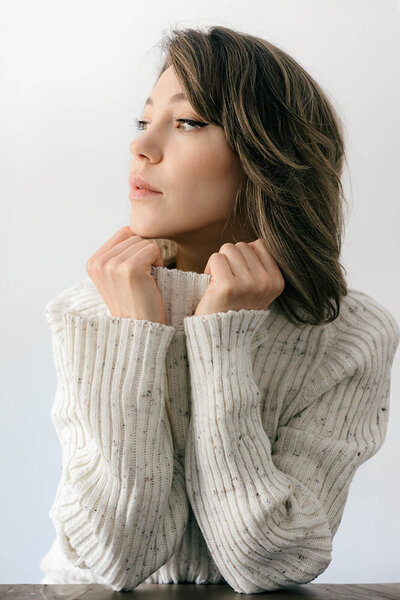 portrait of a woman wearing a cozy sweater color beige. She is holding the turtleneck and looking outside. 