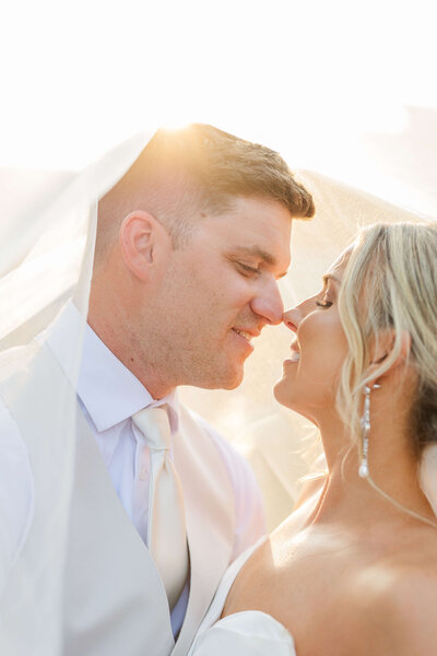 A bride and groom stand under her veil as it's flipped over head. They are smiling and the sun is coming through.