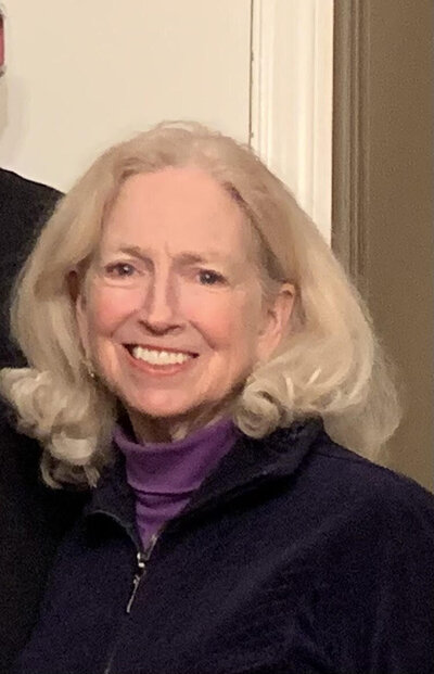 Smiling senior woman with long blonde hair, wearing a purple turtleneck and navy zip-up jacket, standing indoors against a neutral background.
