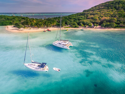 Two sailboats float on clear turquoise waters near a tropical island, perfect for Caribbean getaways, with a sandy beach and lush green vegetation. A small hill rises on the right, and the sky is a soft pink, suggesting sunset or sunrise. A small dinghy is tied to one boat.