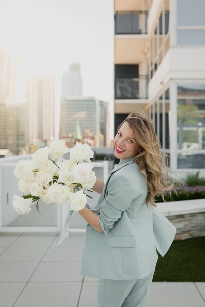 Beautiful woman with light skin with long blond hair is walking on a rooftop in Chicago with the skyline in the background. She is wearing a mint green suit, holding a large bouquet of white flowers, and turning to laugh at the camera.