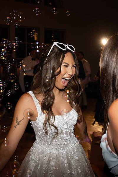 Bride in a white floral gown and white sunglasses laughing joyfully as bubbles float around her on the dance floor.