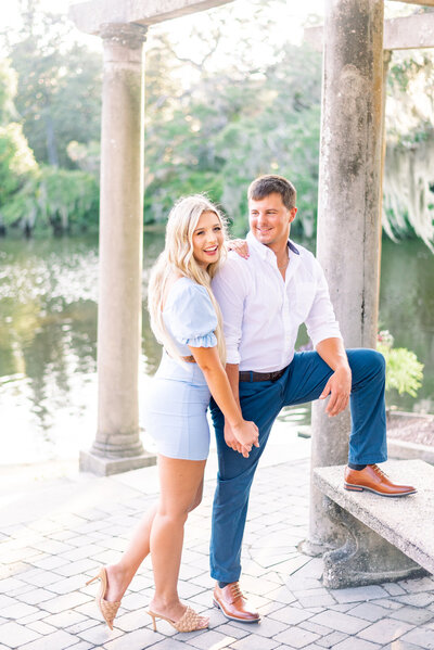 A man and a woman pose for a photo in front of a lake. The man is looking at the woman and the woman is looking at the camera, both similing.