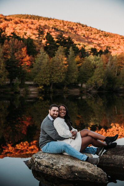 man and woman embracing while sitting on large rock next to a lake