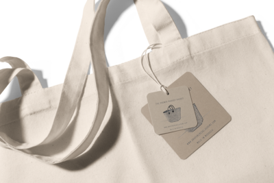 Close-up of a beige canvas tote bag with two tags attached to one of the handles via string, displaying the Sweet Pea Ink Creative logo and website information, beautifully showcasing their brand identity.
