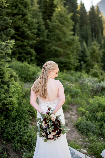 Bride with her back to the camera holding a bouquet behind her back in the mountains