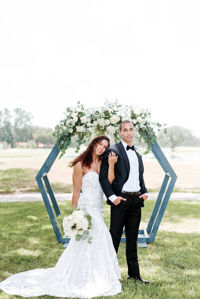 bride holding onto her grooms arm up to  his shoulder and leaning into him as she stands in front of a ceremony arch and holds a white wedding bouquet in her other hand.