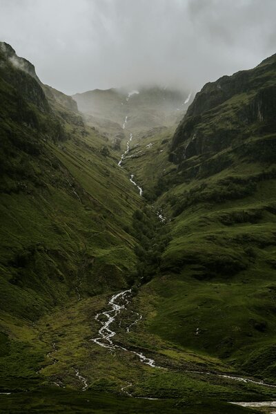 Evocative image of a lush green mountain, where water cascades down between two peaks, shrouded in mist and clouds. The moody atmosphere enhances the dramatic beauty of this natural scene.