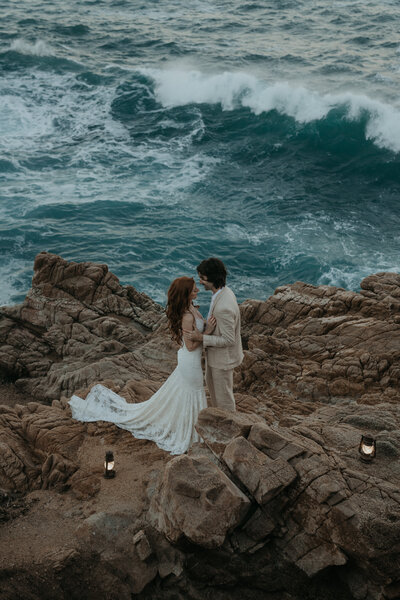 Bride and groom standing on rocks with ocean in the background there are lanterns on the rocks