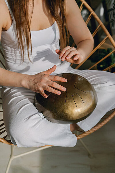 Femme habillée en blanc assise en tailleur dans une chaise et faisant des sons avec un instrument en bronze.