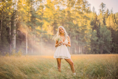 blonde senior girl with long curly hair standing in a grassy field with trees in the background in glacier national park montana