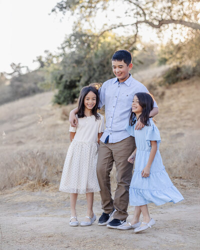 Older siblings playfully pose and hug with elder brother having his arms around his younger twin sisters during a golden hour photoshoot at San Jose
