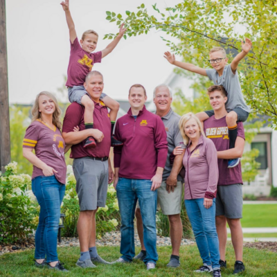Extended family photo outdoors with grandparents, parents, and kids all smiling and cheering together in matching sports team shirts.